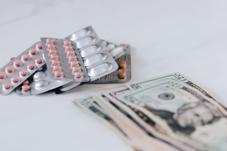 Multiple blister packs of pink and white pills arranged on a white surface alongside a small stack of US dollar bills, with some pills scattered around. The blister packs contain round tablets in vari