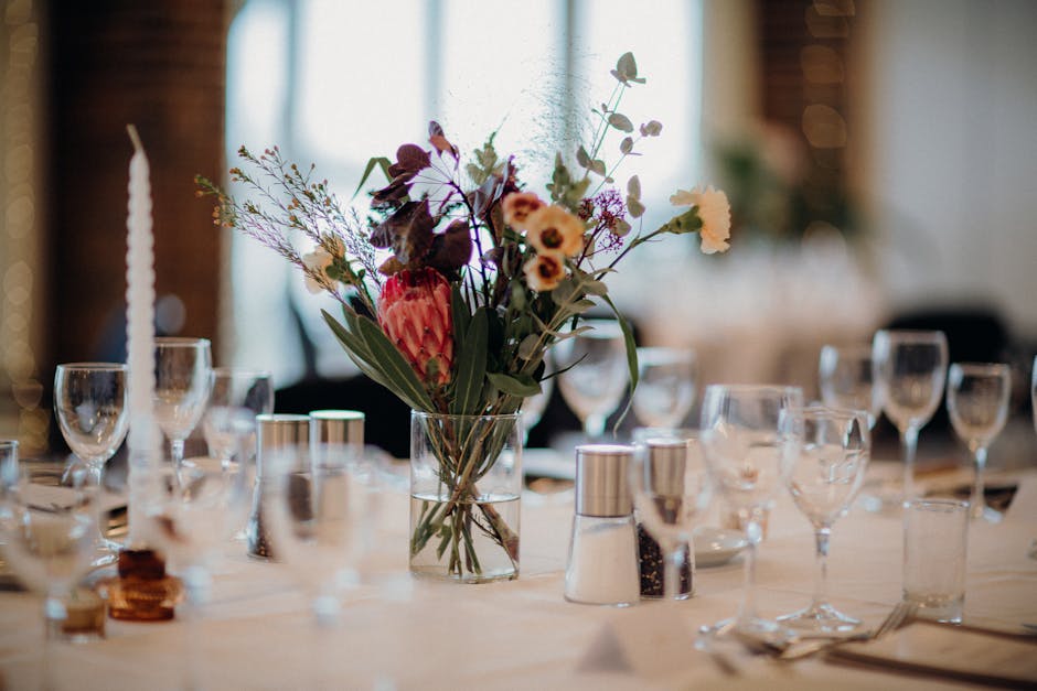 A floral arrangement featuring a variety of fresh flowers in a clear glass vase, placed on a banquet table set for a special occasion. The bouquet includes purple and white flowers, with some pink and