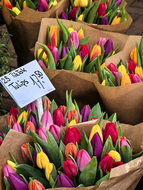 A collection of fresh tulips with vibrant pink, yellow, purple, and orange petals, arranged neatly in brown kraft paper wrapping. The flowers are displayed outdoors on a wooden surface, with some tuli