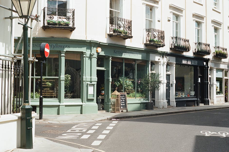 A charming flower shop with a pastel green façade located on a city street corner, featuring large glass windows showcasing floral arrangements inside. The shop has a black chalkboard sign outside ad