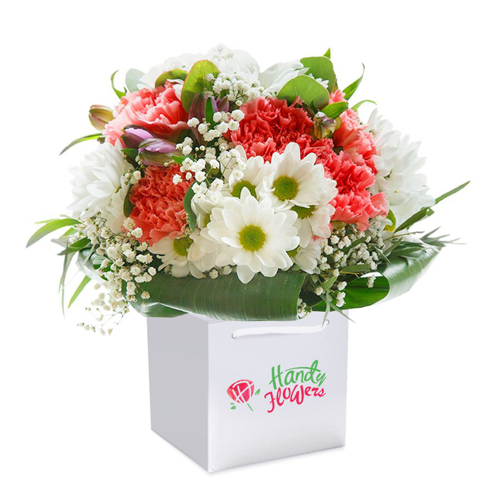A striking and beautifully composed floral bouquet, inspired by the neighbourhood charm of Gipsy Hill, stands upright in an elegant white square gift bag, set against an immaculate white backdrop. The arrangement's heart beats with lush coral and blush-pink carnations-each bloom a flourish of ruffled softness and depth, reminiscent of flower stalls on Westow Hill in the spring. Nestled between these textured blossoms are pristine white daisies, accented with striking lime-green centres, radiating happiness and youthfulness that evoke leisurely afternoons in nearby parks. Interspersed throughout, airy sprigs of baby's breath offer a dreamy, cloudlike lightness, while several purple-tinted buds-perhaps unopened alstroemeria-peek from the left, adding a subtle intrigue that draws the eye, much like a secret corner garden. Encircling all, a collar of broad, glossy, deep-green leaves wraps the blooms in a protective embrace, grounding the pastel palette and giving shape to the composition. The gift bag, with its simple ribbon handles, is as sophisticated as it is practical-ready for hand delivery and evoking the promise of thoughtful gifting on College Road. Though branding is absent, the arrangement's freshness and elegance are clear, making this the ideal gift for birthdays, anniversaries, or any treasured moment that deserves a burst of colour and heartfelt emotion. This image captures not only the high quality of local floristry but also the authentic spirit and vibrancy of the Gipsy Hill community.