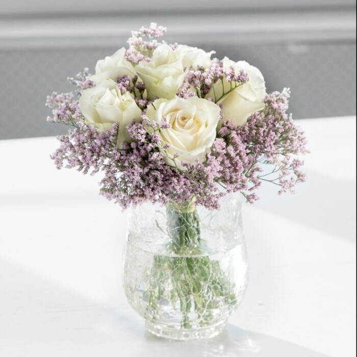 A carefully composed bouquet of white roses and lavender-tinted statice is captured in soft, natural light, the kind that often pours through bay windows along Gipsy Hill on a clear afternoon. The flowers are gathered densely into a rounded, symmetrical arrangement that feels both deliberate and effortlessly graceful. At its heart, around a dozen creamy white roses open in varying stages, some still cupped and shy, others gently unfurled to reveal intricate whorls of petals. Their colour is a warm, almost ivory white, with subtle shading near the centres that gives them depth and dimension. Framing and threading between the roses, feathery sprays of pale lilac statice create a delicate haze, their many small blossoms forming a soft, cloud-like border. This contrast between the lush, velvety rose petals and the fine, papery texture of the statice gives the bouquet a layered, tactile quality you can almost feel. The stems are gathered neatly in a tall, cylindrical glass vase, clearly visible through the transparent sides, where they are anchored in sparkling water that catches tiny reflections of green. The vase stands on a smooth white surface, possibly a kitchen island or side table in a Crystal Palace or West Norwood flat, its simplicity allowing the flowers to take full focus. The background is gently blurred, suggesting a calm indoor setting with plenty of daylight, which brings out the serene colour palette of cream, white, and soft lavender. The overall impression is one of poised, modern elegance: a bouquet suited to marking a quiet celebration, sending condolences with subtle beauty, or welcoming someone into a new home in the Gipsy Hill area with a sense of peace and thoughtful care.